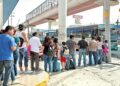 Crowd of Commuters Waiting at a Bus Stop As a Blue-green City Bus Pulls Up Nearby Under an Overpass. - La Sultana Informa