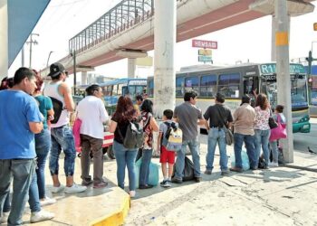 Crowd of Commuters Waiting at a Bus Stop As a Blue-green City Bus Pulls Up Nearby Under an Overpass. - La Sultana Informa