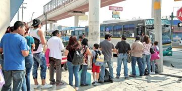 Crowd of Commuters Waiting at a Bus Stop As a Blue-green City Bus Pulls Up Nearby Under an Overpass. - La Sultana Informa