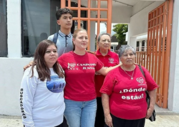 Group of Five People Posing Outside a Building with Orange Gates; Two Women Wear Red Shirts, One in a White Long-sleeve Shirt with 'buscadores' on the Sleeve, and a Teen Behind Them. - La Sultana Informa