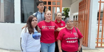 Group of Five People Posing Outside a Building with Orange Gates; Two Women Wear Red Shirts, One in a White Long-sleeve Shirt with 'buscadores' on the Sleeve, and a Teen Behind Them. - La Sultana Informa