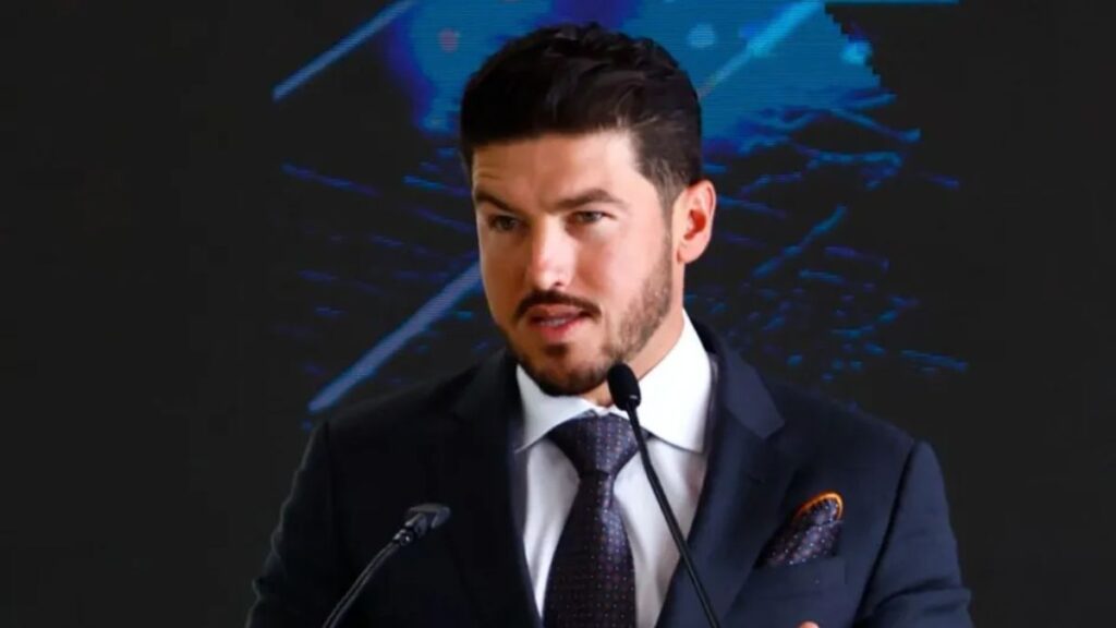 Samuel-garcia-guerra-sucia - La Sultana Informa Man in a Dark Suit and Tie Speaks into Two Microphones at a Podium, with a Blue Abstract Backdrop Behind Him. - La Sultana Informa